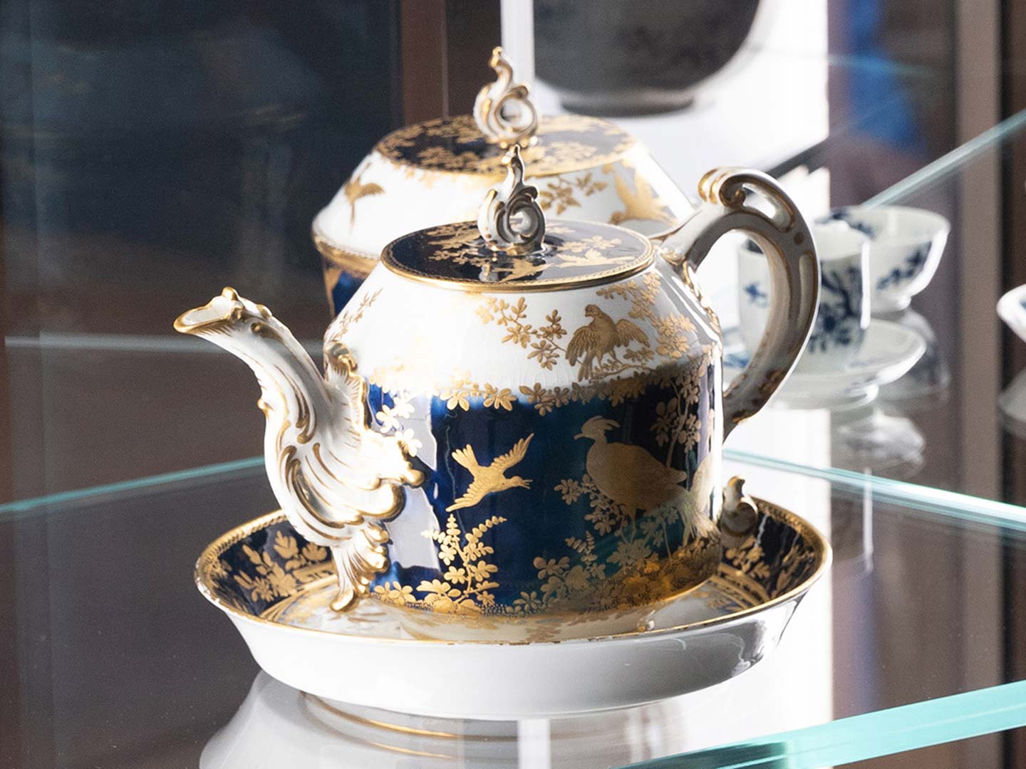 A blue, white, and gold porcelain teapot with chinese-inspired bird shapes painted on its side, sitting in a glass case